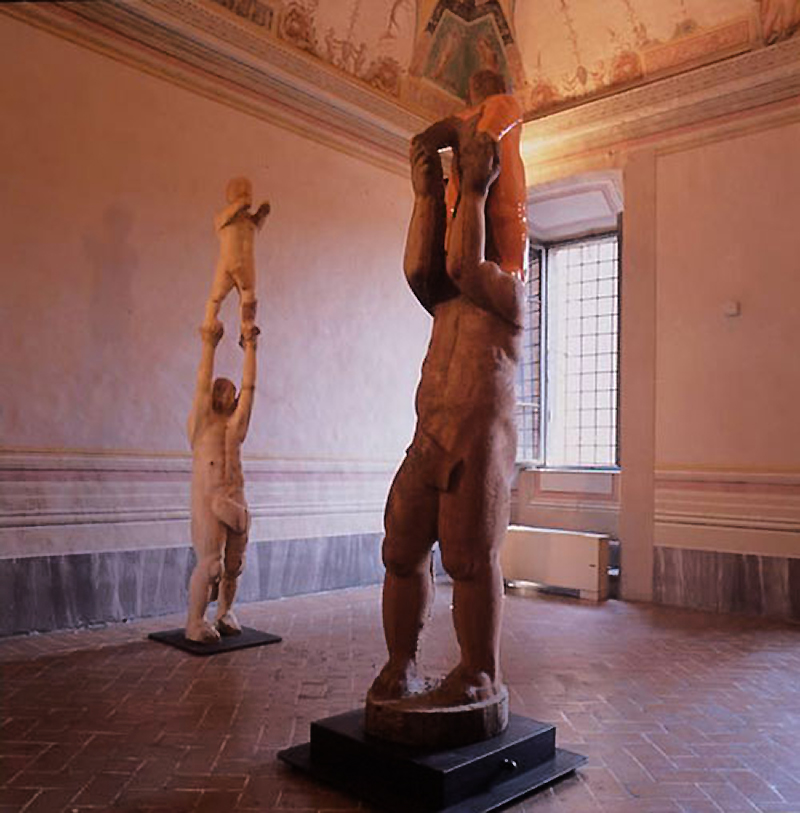 Due sculture figurative in legno ciascuna rappresenta una piccola figura sulle spalle dell'altra esposte nella Sala del Concerto di Palazzo della Corgna a Città della Pieve in provincia di Perugia